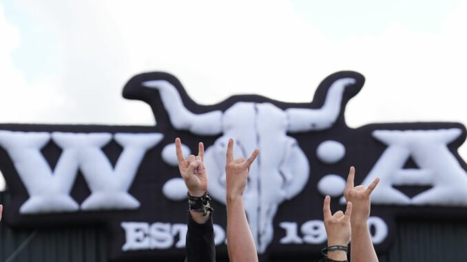 Wacken Open Air Metal-Fans zeigen vor dem Wacken Open Air-Logo die „Pommesgabel“, den Metal-Gruß, auf dem teilweise matschigen Festivalgelände des Wacken Open Airs (WOA).