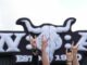 Metal-Fans zeigen vor dem Wacken Open Air-Logo die „Pommesgabel“, den Metal-Gruß, auf dem teilweise matschigen Festivalgelände des Wacken Open Airs (WOA).